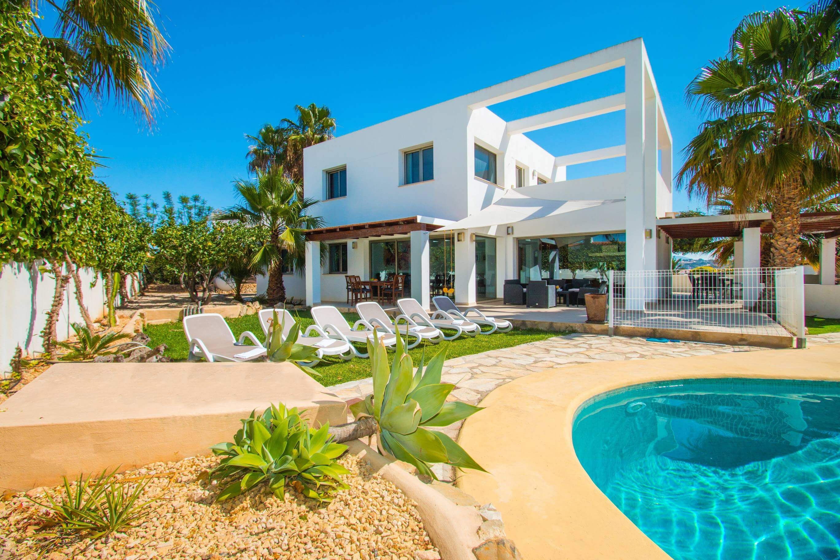 Una casa moderna de dos pisos amb una piscina, espai d'estar a l'aire lliure i palmeres.