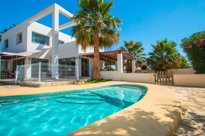 A modern two-story house with a swimming pool, palm trees, and a clear blue sky.