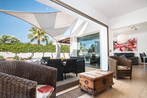 A modern outdoor lounge area with wicker furniture, a wooden chest, and a large umbrella providing shade.