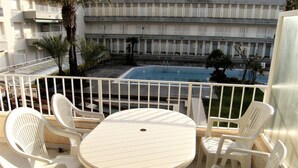 Un balcón con una mesa redonda y sillas de plástico blancas, con vista a una piscina y un edificio con balcones.