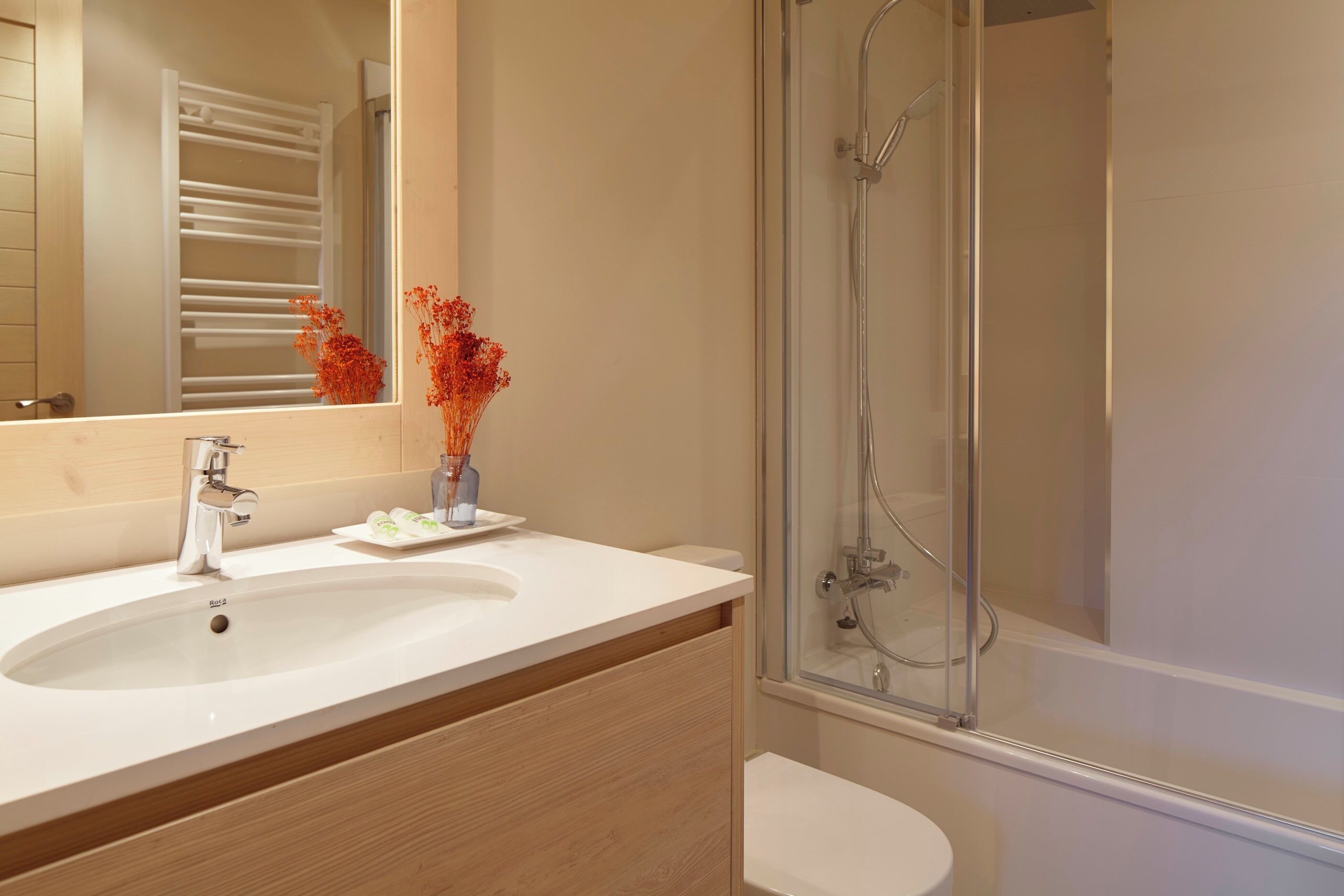 Une salle de bain moderne avec un grand miroir, un lavabo, une douche à parois de verre et un petit vase avec des fleurs.
