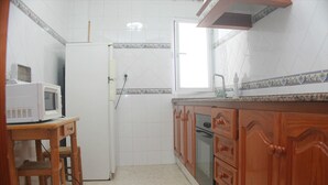 A compact kitchen with white tiled walls, a microwave on a wooden table, wooden cabinets, and a window.