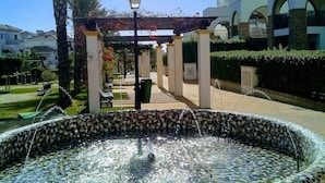 Ein Brunnen mit Mosaikfliesen, eine Palme und im Hintergrund ein Gebäude mit Bögen.