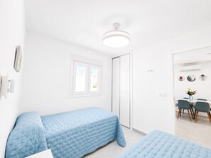 Un dormitorio con dos camas individuales, una ventana, una puerta, una lámpara en el techo y una zona de comedor visible a través de una puerta abierta.