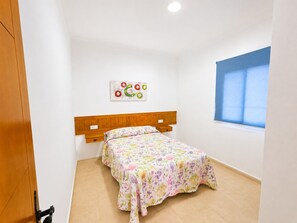 Ein Einzelbett mit Holz-Kopfstück in einem Zimmer mit weißen Wänden und einem Fenster mit blauen Jalousien.