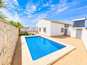 A swimming pool with a stone wall on one side and a white building with a tiled roof in the background. - Chalet Playa Roche . (Conil de la Frontera)