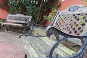 Um banco de madeira com apoios de braço de metal e encosto treliçado, em frente a uma parede vermelha e vegetação.