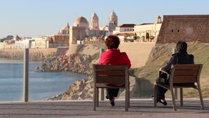 Dos personas sentadas en bancos con vistas a una zona costera y edificios históricos al fondo.