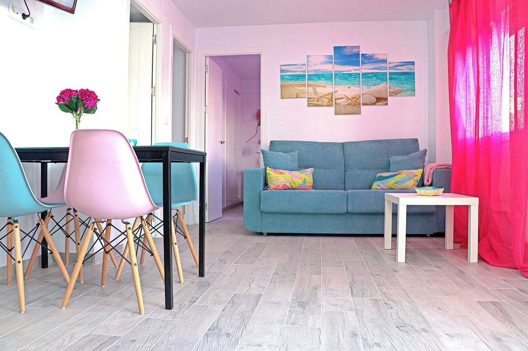 A living room with a blue sofa, teal and pink chairs, a white table, and a painting of a beach scene.