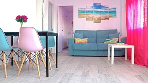 Un salón con un sofá azul, sillas verde azulado y rosa, una mesa blanca y un cuadro de una playa.