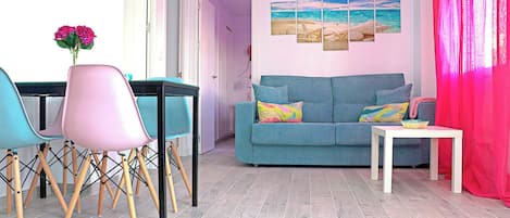 A living room with a blue sofa, teal and pink chairs, a white table, and a painting of a beach scene.