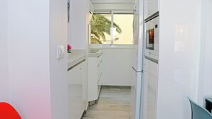 Un pasillo estrecho con paredes blancas, una ventana con vistas a palmeras y un refrigerador a la derecha.
