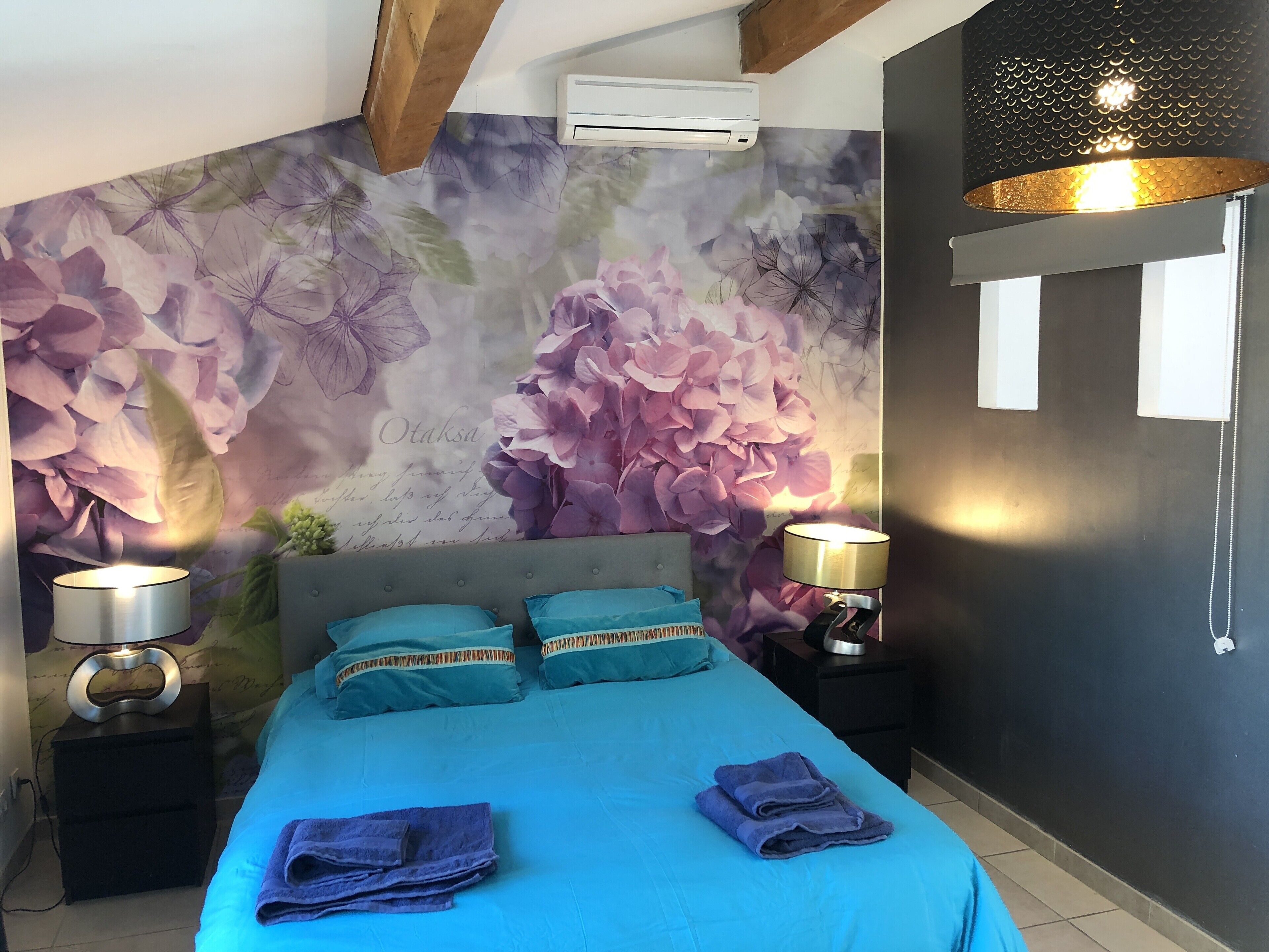 A bedroom with a floral wall mural, a bed with blue bedding, two bedside lamps, and a modern air conditioning unit.