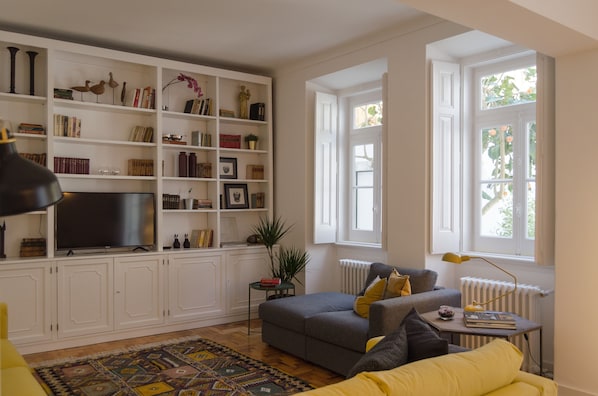 A living room with a bookshelf, a grey sofa, a yellow sofa, a patterned rug, and a window with a view of greenery.