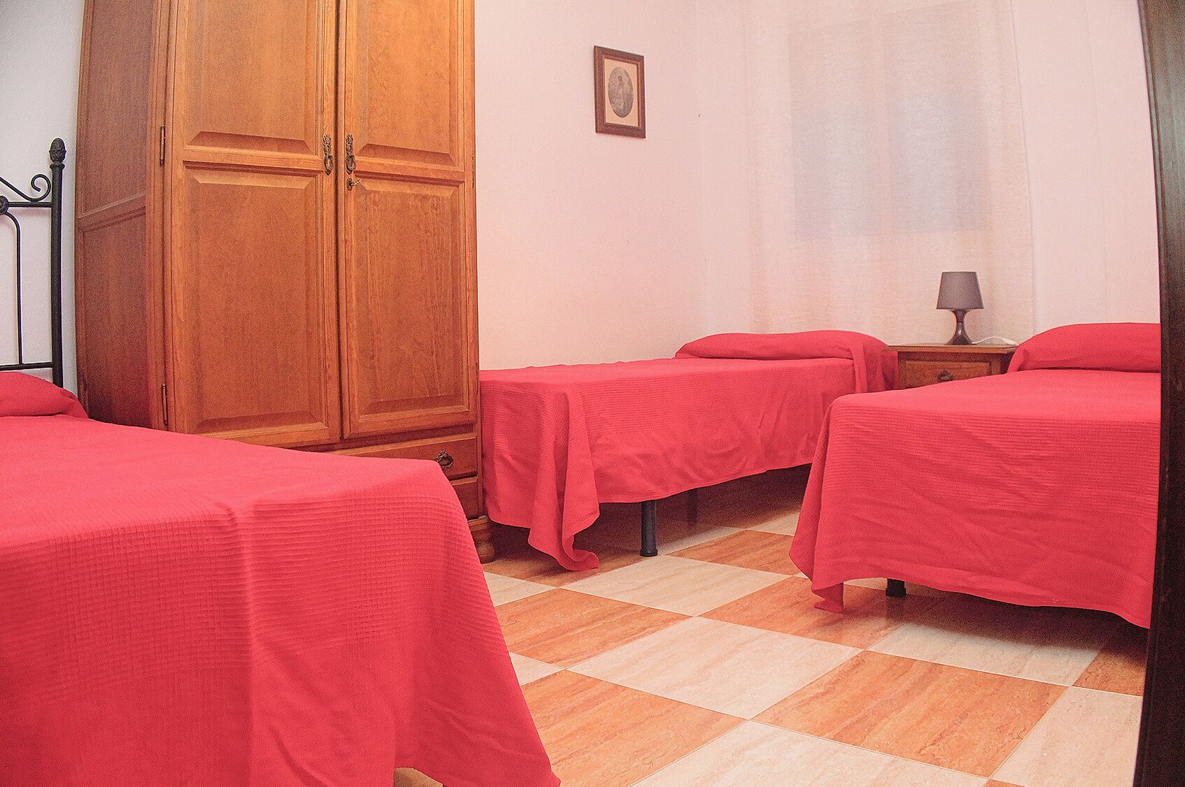 A room with two beds, a wooden wardrobe, a small bedside table with a lamp, and a checkered floor.