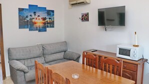 Un espacio habitable compacto con un sofá gris, una mesa de comedor de madera y sillas. Un televisor de pantalla plana está montado en la pared sobre la mesa.