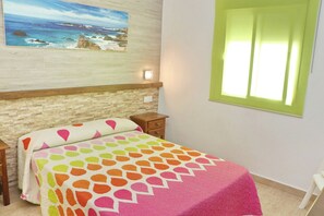 Une chambre à coucher comprenant un lit, une table de chevet, une lampe fixée au mur, une fenêtre avec des stores verts et un tableau représentant un paysage côtier.
