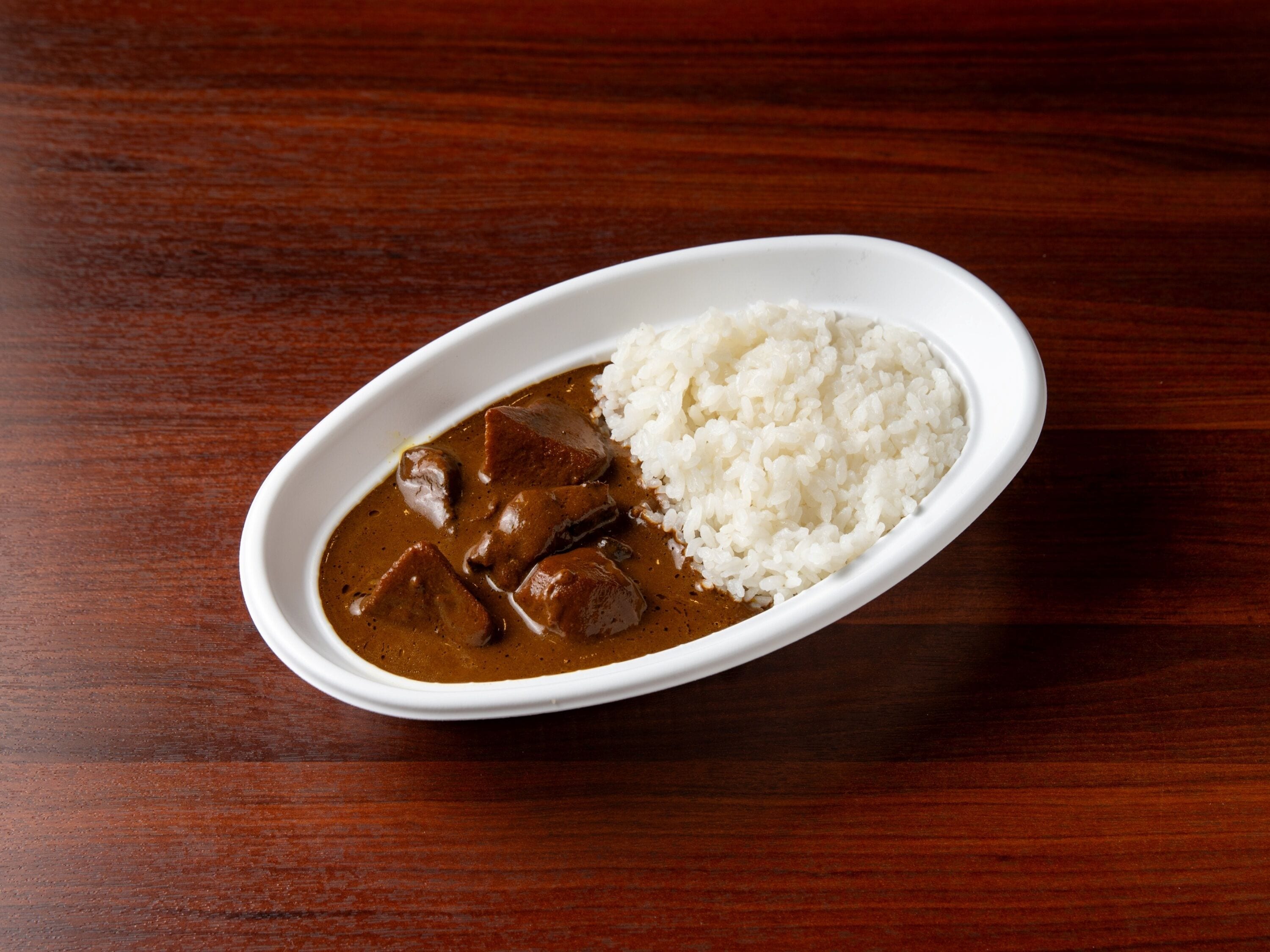 A white oval dish containing a brown curry with chunks of meat and white rice on a wooden table.