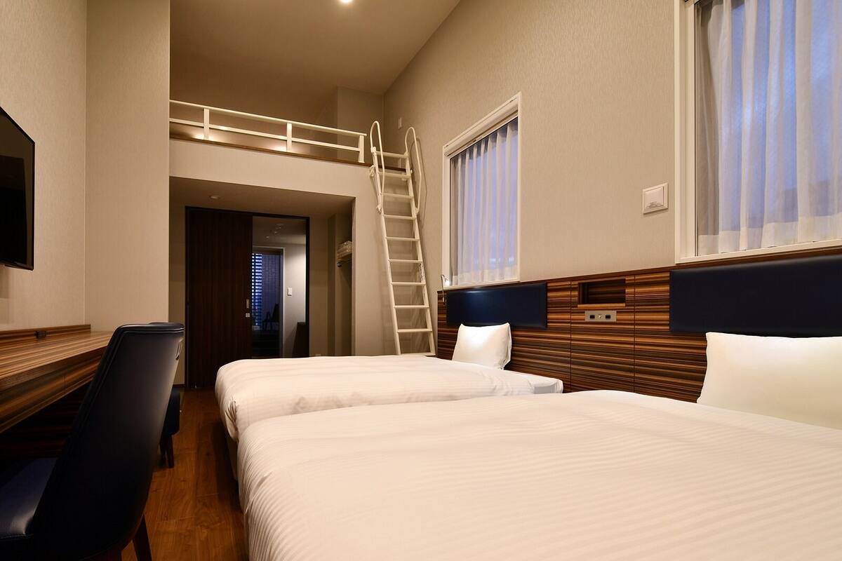 A hotel room with two beds, a desk, a chair, a TV, and a window with curtains.