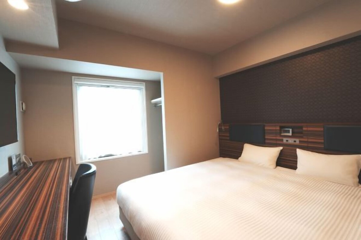 Habitació d'hotel amb llit, escriptori, finestra i televisió penjada a la paret.