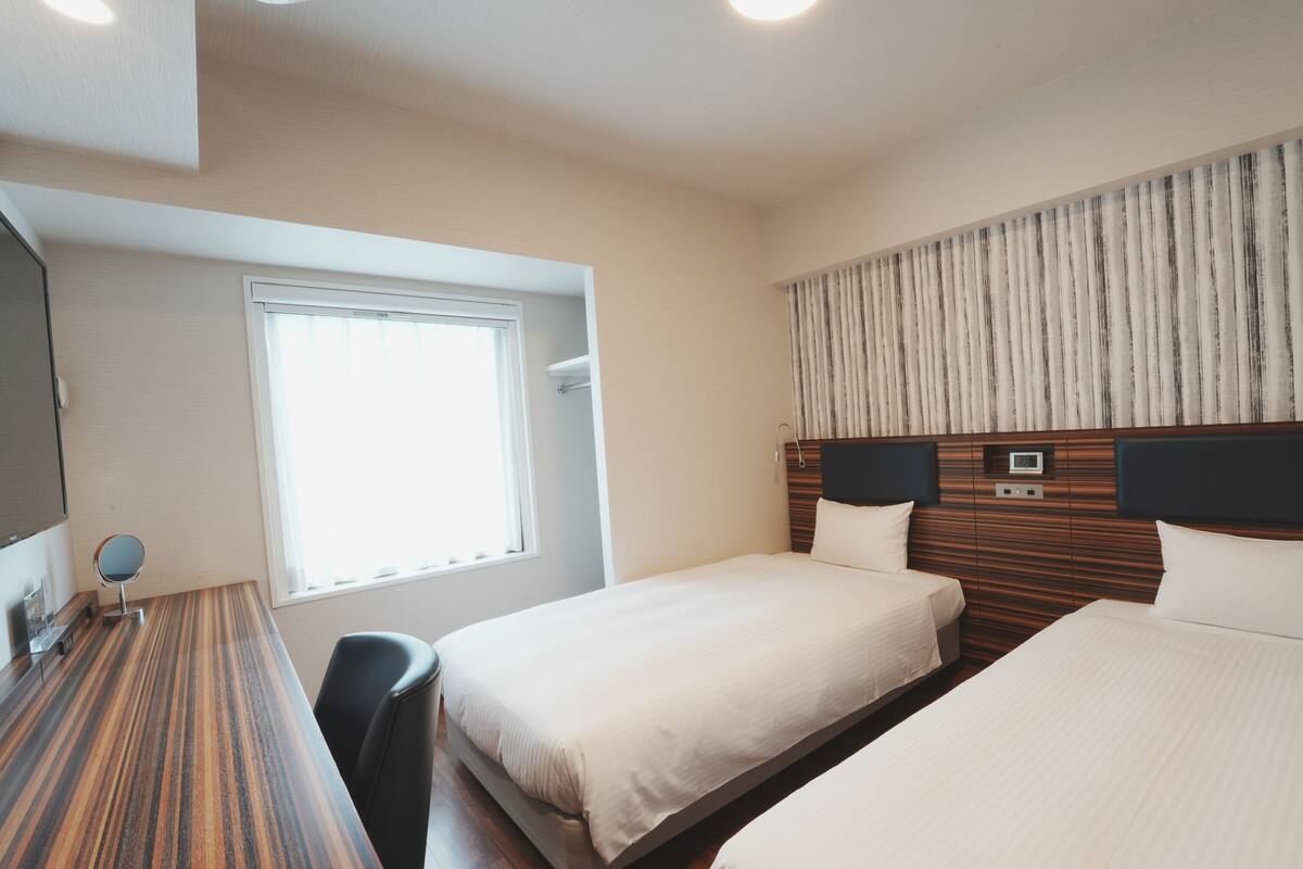 A hotel room with two beds, a desk, a chair, a mirror, a window, and a curtain.