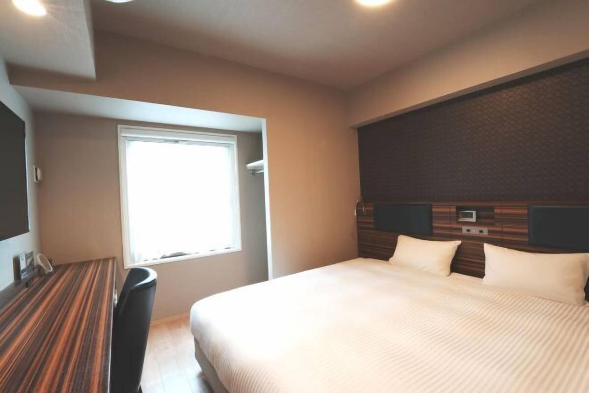 A hotel room with a bed, a desk, a window, and a wall-mounted TV.