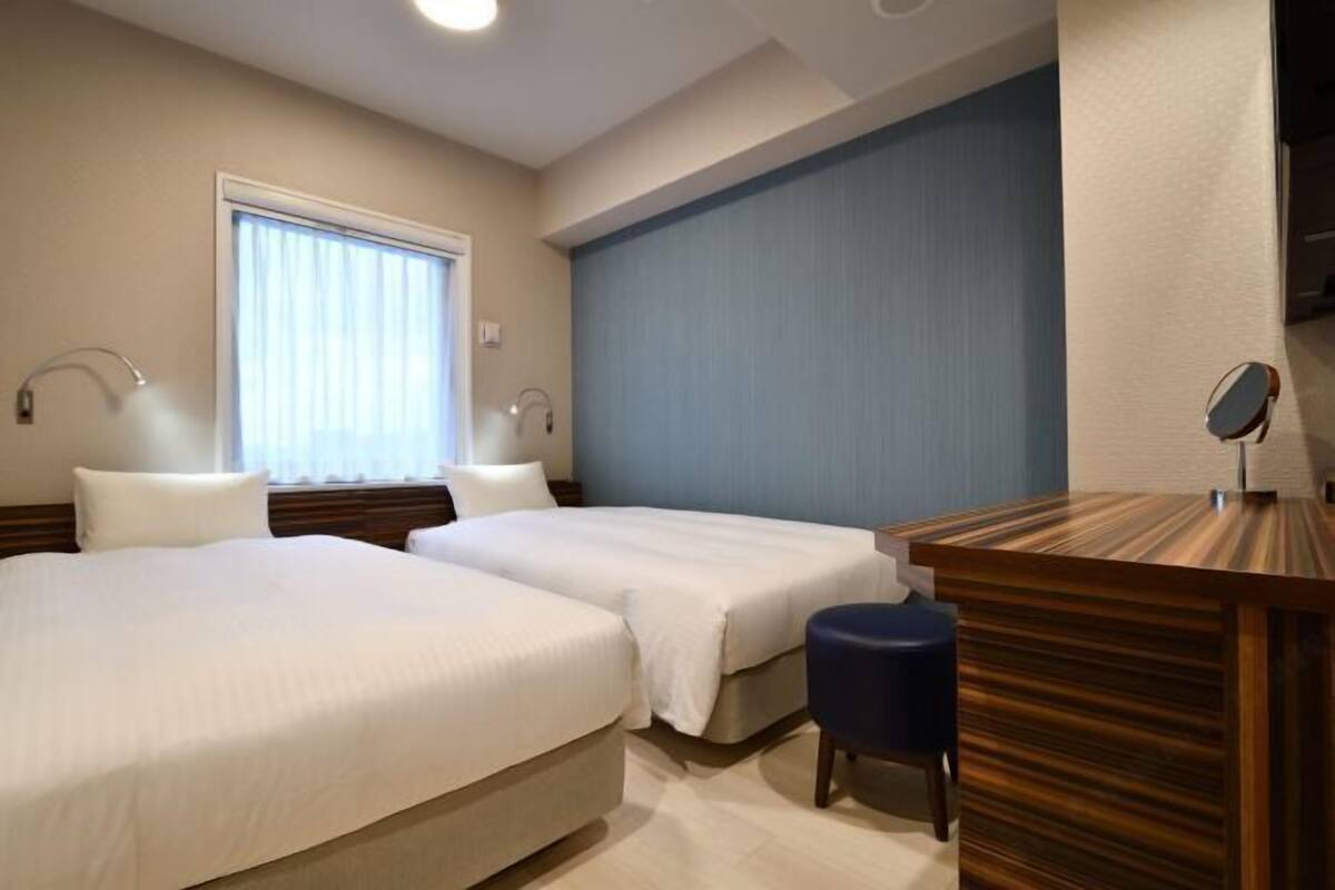 A hotel room with two beds, a desk, a chair, a window with curtains, and a wall-mounted TV.