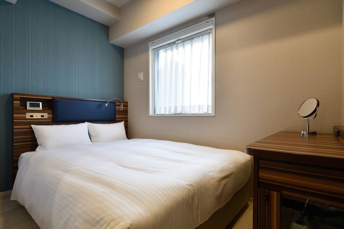 A hotel room with a bed, a bedside table with a mirror, a window with curtains, and a wall clock.