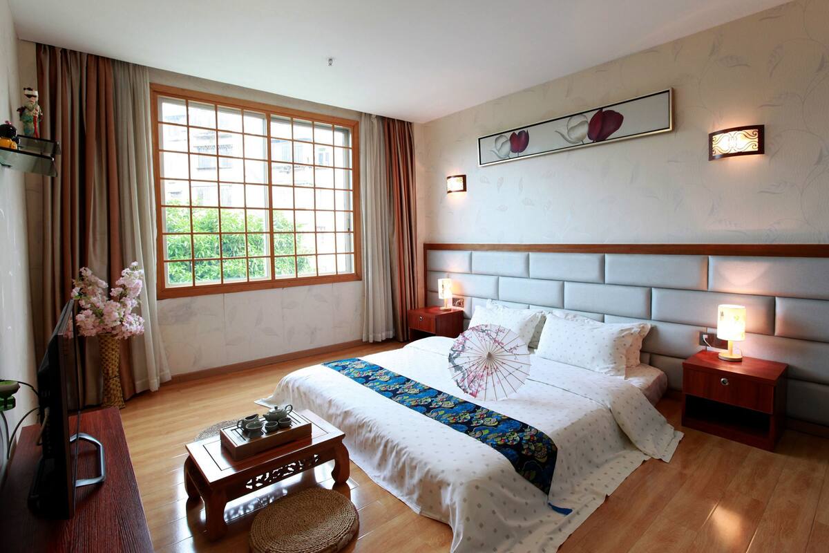 A bedroom with a large window, a bed with a headboard, bedside tables with lamps, a wooden desk, and a decorative vase with flowers.