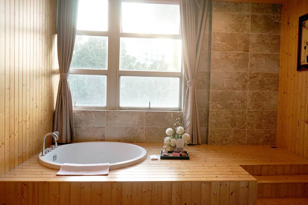 A wooden-floored bathroom with a large bathtub, a window with curtains, and a small table with a vase of flowers.