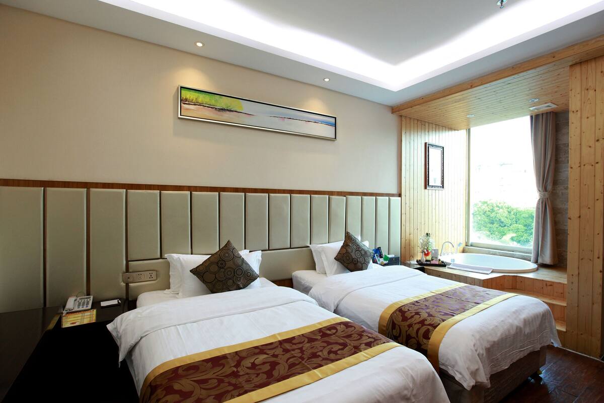 A hotel room with two beds, a large window with curtains, a painting on the wall, and a bedside table with a phone and a lamp.