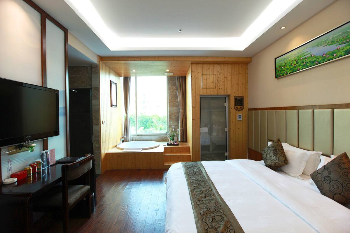 A hotel room with a large bed, a TV, a desk with a chair, a window with a view of greenery, and a shower area.