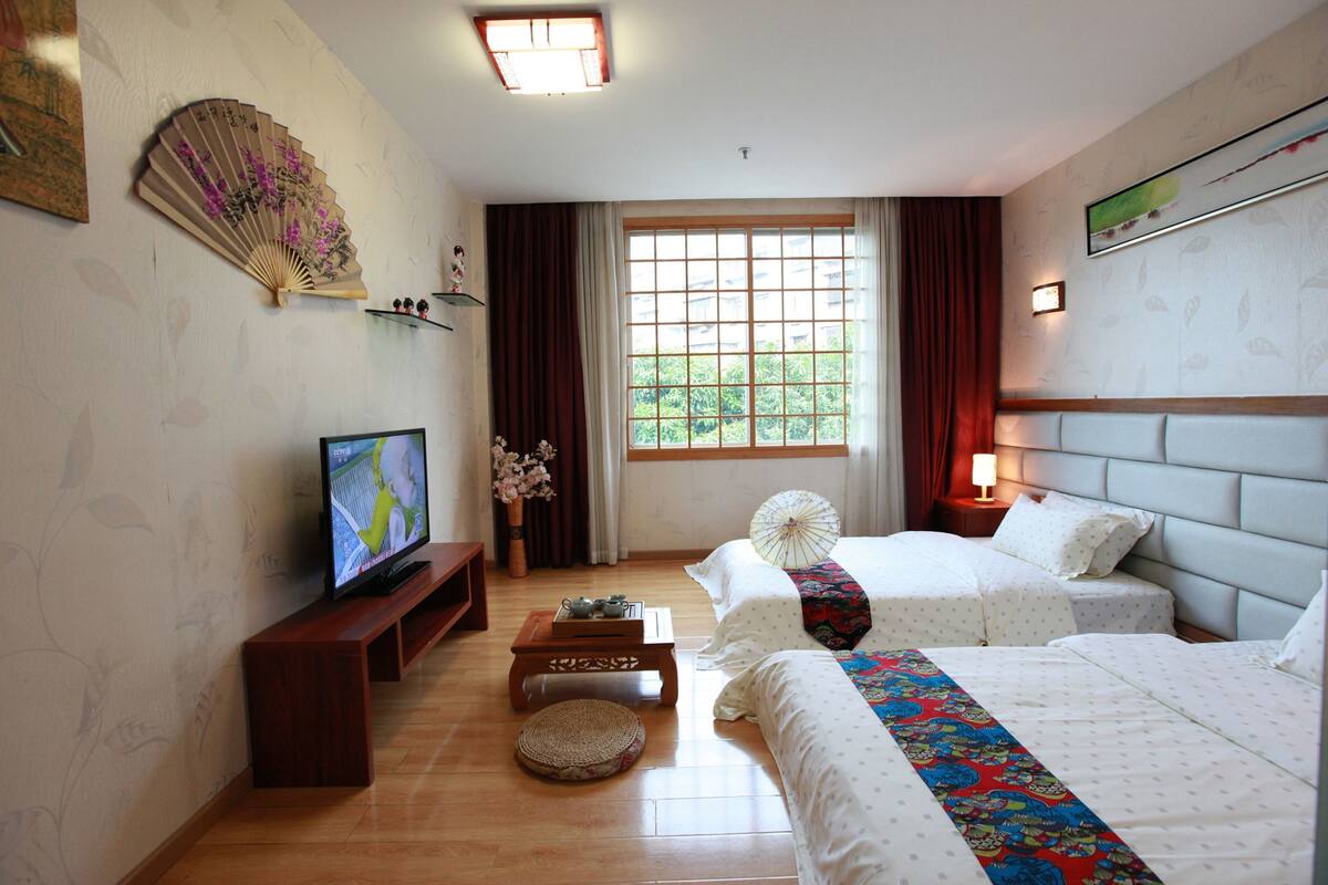 A hotel room with two beds, a TV, a small table, a decorative fan, and a window with curtains.