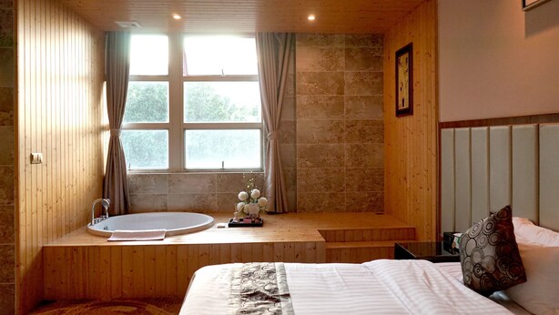 Un dormitorio con piso de madera, una cama con cubrecama blanco, una mesita de noche, un televisor montado en la pared y una ventana con cortinas.