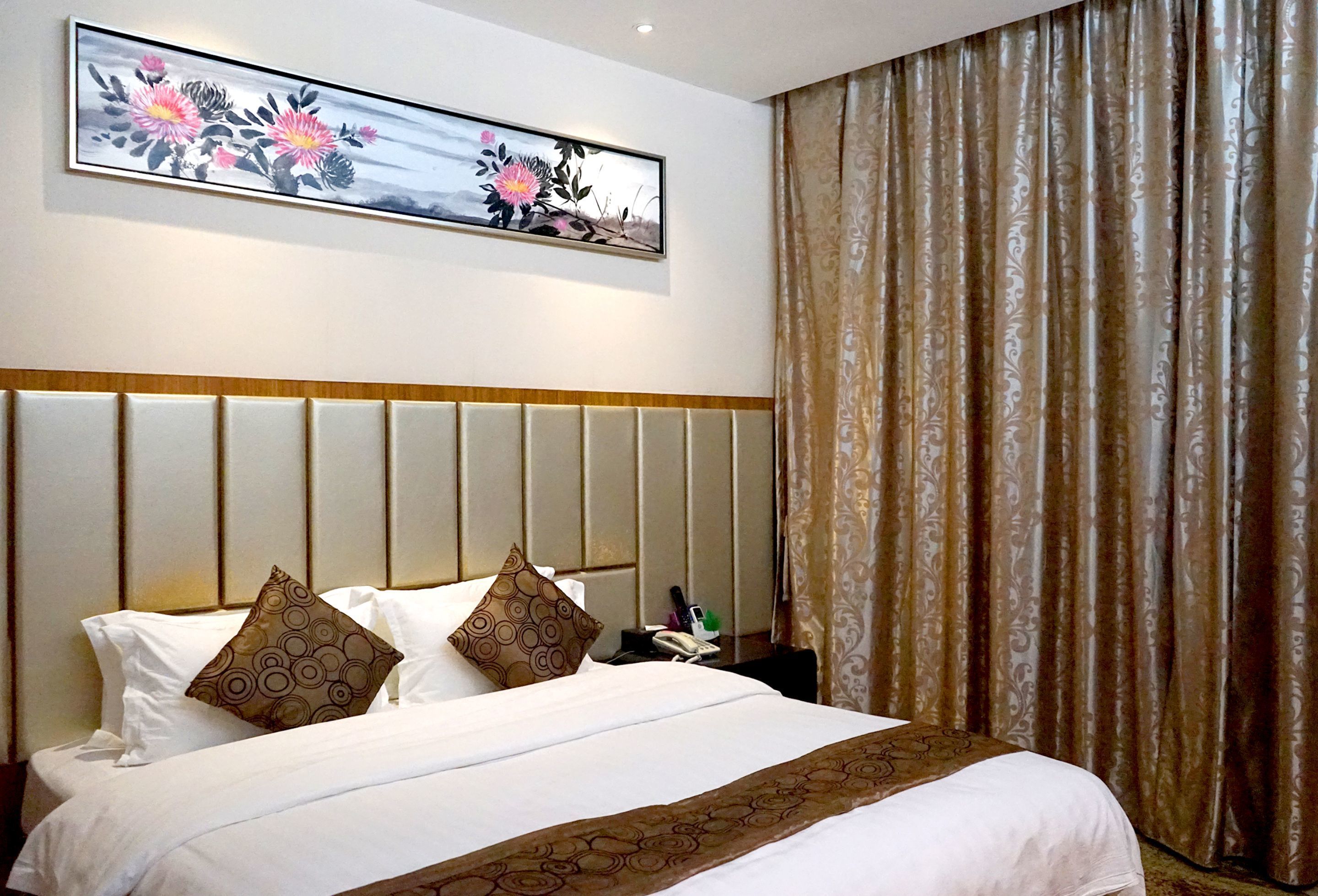 A hotel room with a large bed, a headboard with vertical panels, a painting above the bed, and a curtain on the window.