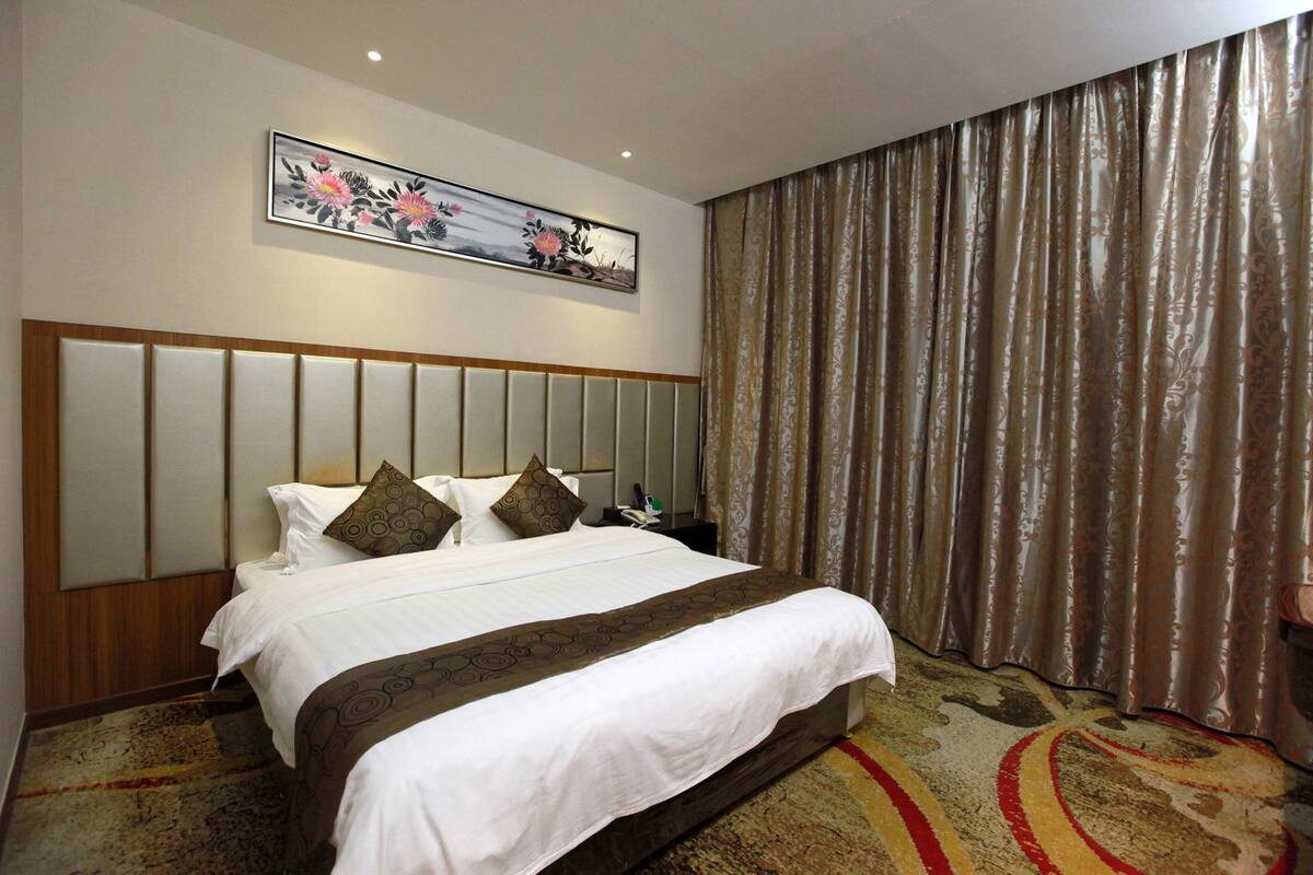 A hotel room with a large bed, a headboard with vertical panels, a decorative painting above the bed, and a patterned carpet.