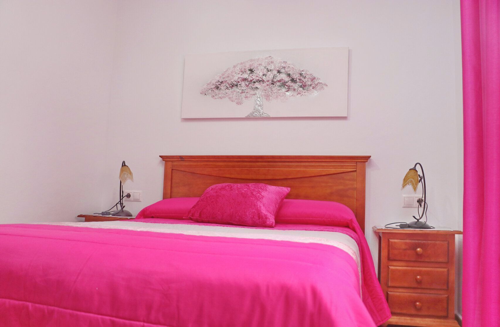 Ein Schlafzimmer mit einem hölzernen Bett, rosa Bettwäsche, einem Nachttisch mit Lampe und einem Wandbild.