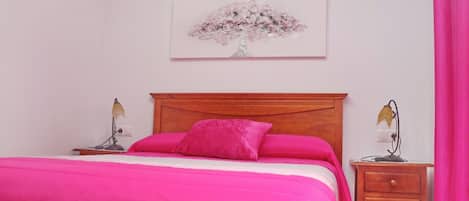 Een slaapkamer met een houten bed, roze beddengoed, een nachtkastje met een lamp en een aan de muur hangend kunstwerk.