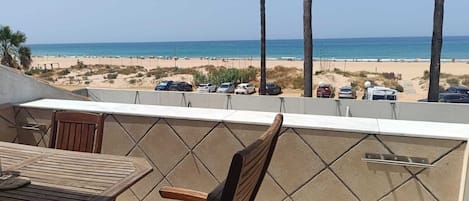 A balcony with a view of the beach, a table, chairs, and a railing.