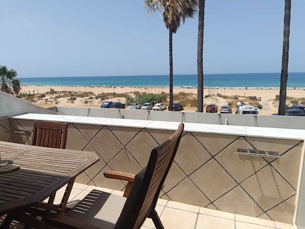 A balcony with a view of the beach, a table, chairs, and a railing.