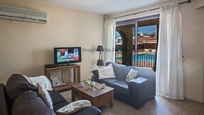 A living room with a sofa, a coffee table, a TV, and a balcony with a pool view. - An apartment that sleeps 4 guests  in 2 bedrooms (Ayia Napa)
