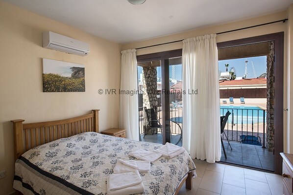 A bedroom with a bed, an air conditioner, a painting, and a view of a pool and beach through a sliding glass door.
