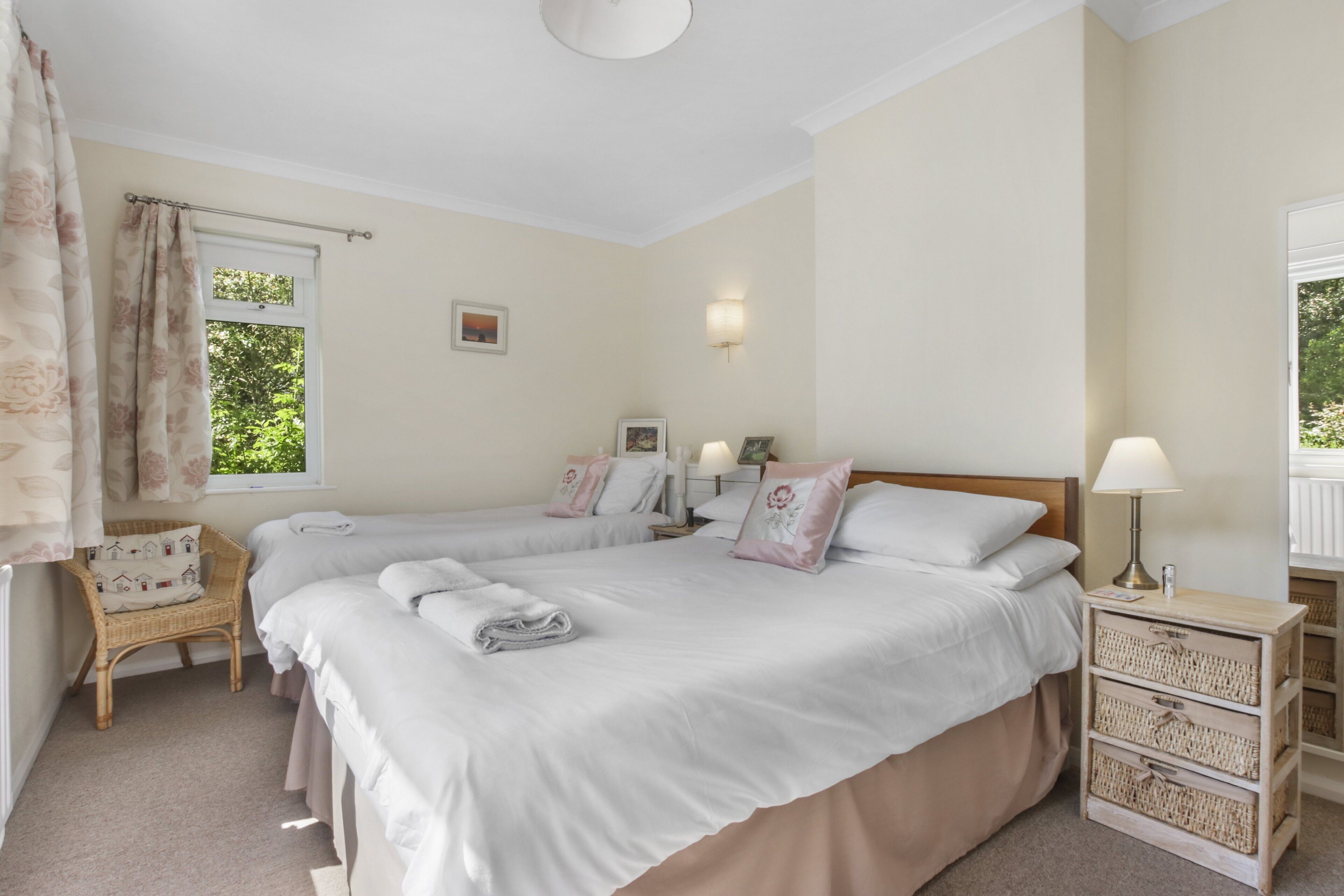 A bedroom with two beds, a wicker chair, a bedside table with a lamp, and a window with curtains.