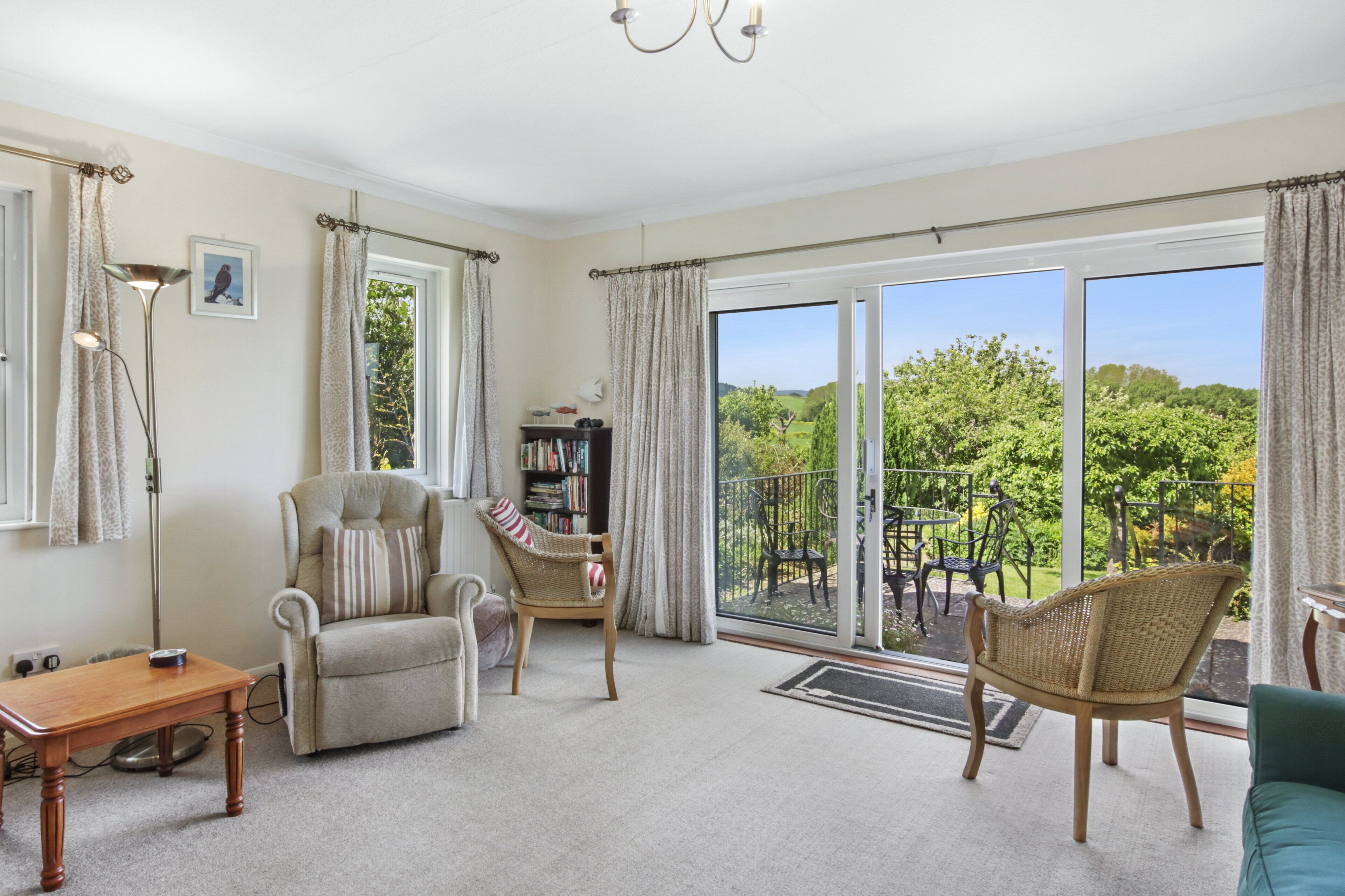 A spacious living room with a large sliding glass door leading to a balcony, a comfortable sofa, a chair, a small table, and a bookshelf.