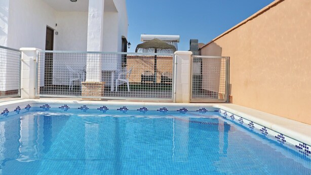 A swimming pool with tiled edges, surrounded by a white building with a beige wall, and metal gates.
