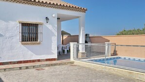 A white building with a tiled roof, a swimming pool, and a covered patio area with chairs.