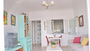A living room with a white sofa, a table, a TV, a cabinet, and a window with curtains. - Apartment in Conil amazing beach views (CONIL DE LA FRONTERA)
