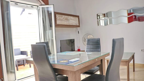 A dining area with a glass-top table, four chairs, a side table, a framed artwork, a wall-mounted mirror, and a sliding glass door leading to an outdoor seating area.