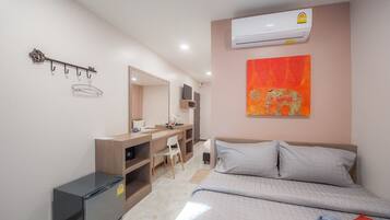 A hotel room with a bed, a desk with a chair, a wardrobe, a mirror, an air conditioner, and a wall-mounted artwork.