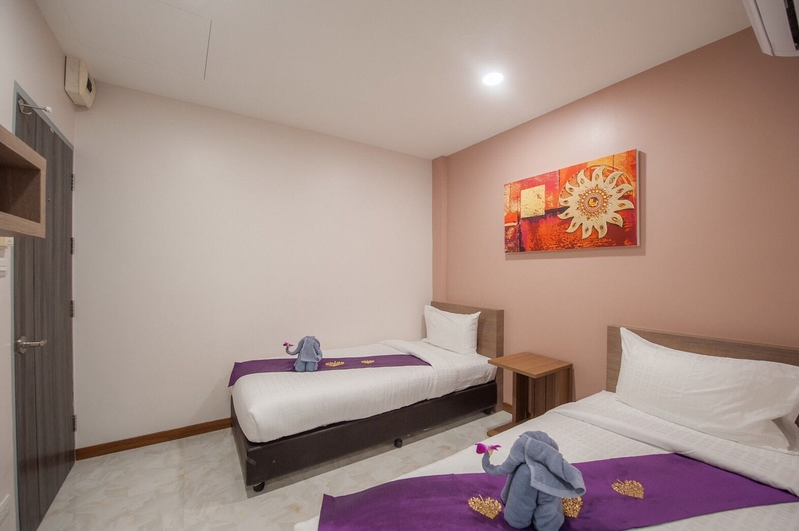 A hotel room with two beds, a small wooden nightstand, a wall-mounted air conditioner, and a colorful abstract painting.